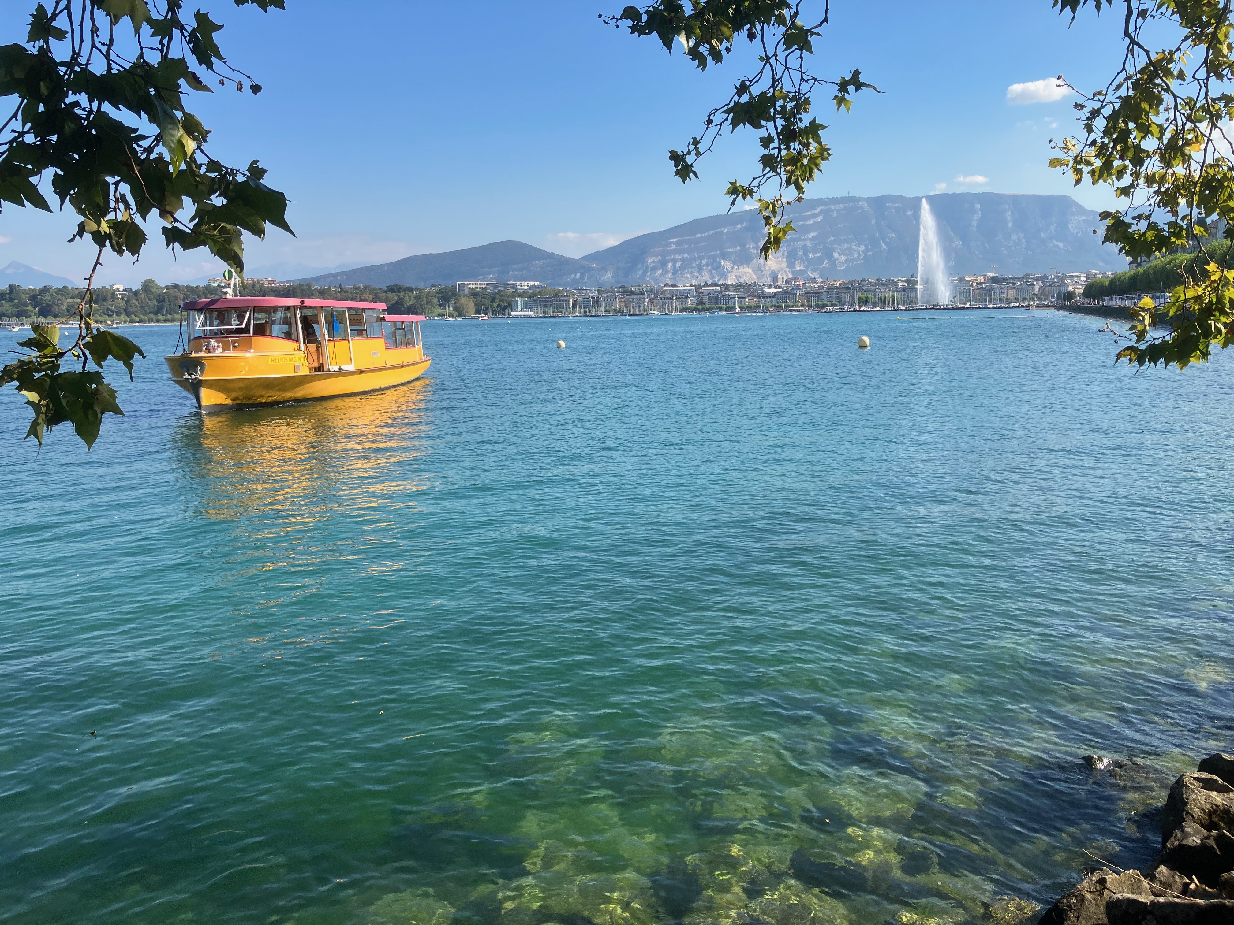 Sunny day with the blue water of Lake Geneva and a yellow boat on it. In the background is the famous Jet d'Eau fountain and the city of Geneva.
